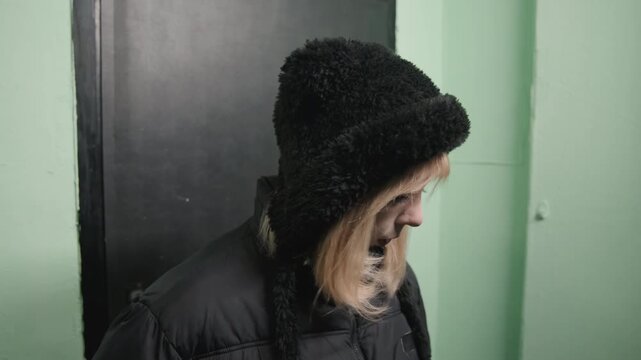 Young white woman near black door hair tucked under earflap hat, turning head with thoughtful gaze, subtle fidgeting and held emotion, cool corridor light, moment of decision, commuter or creative