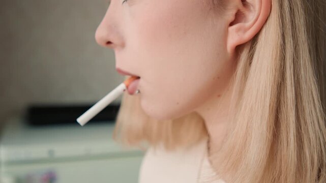 Woman with cigarette pauses peacefully. Calm Caucasian woman with tattoos relaxes by soft light. Tranquil woman with subtle makeup and piercings holds cigarette in gentle kitchen ambiance