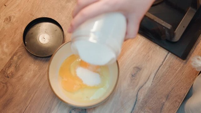 Pouring sugar into cracked eggs bowl, home baker hands tilting jar over yolks on wooden countertop, granulated sugar forming mound in bowl, warm natural light, cozy rustic baking scene preparing
