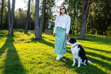 Naklejka premium Young woman with a black and white dog standing in a sunlit forest meadow during summer.