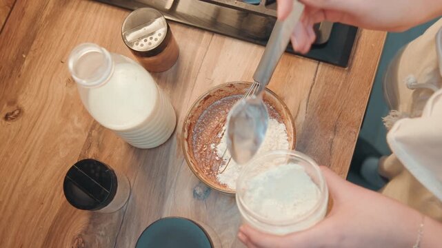 Adding flour to chocolate batter, overhead shot shows milk bottle, whisk and jar on wooden counter, hands fold dry ingredients into cocoa base, bright natural light, tutorial style cooking demo