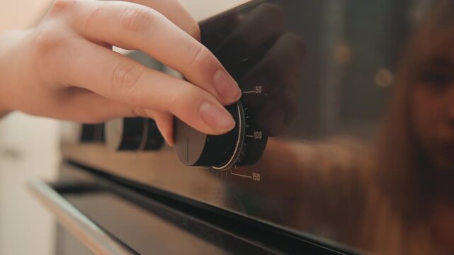 Closeup hand adjusting oven knob, reflective woman watching. Tactile motion on stainless control, warm ambient kitchen light, quiet preparation and focused anticipation before baking.