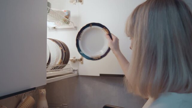 Woman food blogger inspects clean plate at cabinet, close profile shot, checking for spots and shine, thoughtful expression, studio kitchen lighting, preparation for plating content