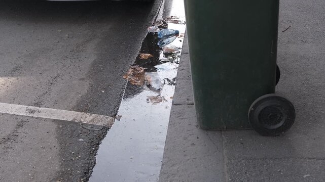 Plastic bottles, autumn leaves, and litter collecting in stagnant water along a street gutter beside a wheelie bin and kerb on an asphalt road in Melbourne, Australia. Discarded single-use plastics