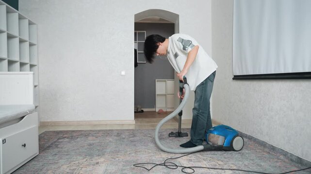 Young man cleans with vacuum. Male student maintains cleanliness by vacuuming his compact room rug. Young Asian man actively uses vacuum cleaner to clean and care for his study carpet