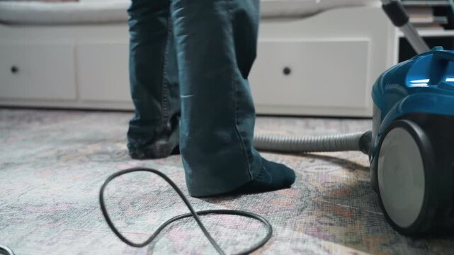 Feet and vacuum cord on carpet. Renter navigates cord and nozzle while quickly vacuuming living room rug, blue canister nearby, socks and jeans visible, dynamic chore with focus on safety