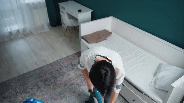 Asian woman vacuuming bedroom rug, topdown view of focused homemaker cleaning near white daybed and desk cordless vacuum gliding across patterned carpet, checking nozzle and moving under bed, serene