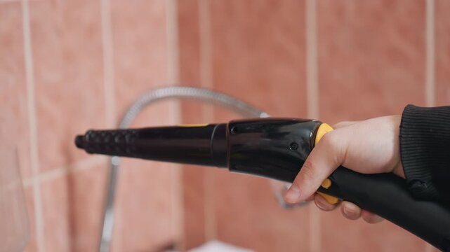 Closeup hand holding steam nozzle over sink by white cleaner, precise grip and trigger control used for grout and tile detail work, intimate equipment shot showing handle, tip, and hose with steady