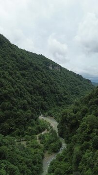 Vertical aerial view of a rocky forested mountain above a river, with a waterfall flowing into the river below in the Egrisi Range, Georgia.