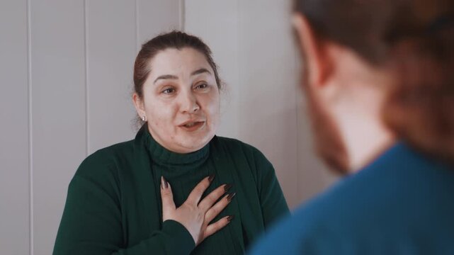 White woman at home speaking to paramedic, hand on chest expressing concern and gratitude while paramedic in blue uniform listens, green sweater, wood paneling background, intimate home medical visit,