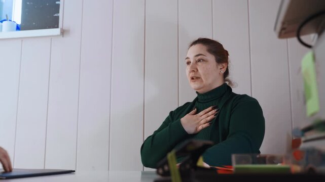 Woman clutching chest at home, seated at small table during urgent medical consultation with responder hand pressed to sternum, anxious expression and shallow breathing, paperwork and small medical