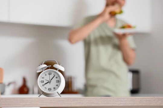 White alarm clock on a kitchen counter with a person eating in the background, time management and healthy eating habits