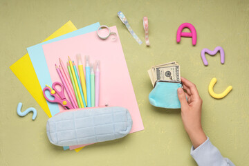 Obraz premium Childs hand holding a coin purse with money next to school supplies and colorful paper on a green background, a childs hand holds a light blue coin purse filled with hundred dollar bills, financial