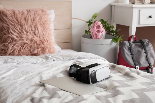 Virtual reality headset and laptop on a bed with a fluffy pink pillow and backpack in a modern bedroom, a fluffy pink pillow leans against a wooden headboard, and a gray and red backpack sits next