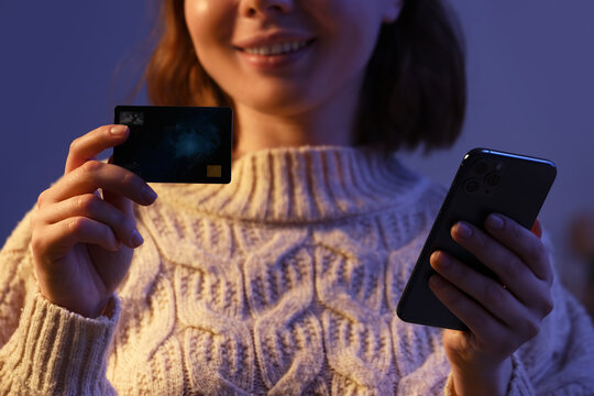Young happy woman with credit card using mobile phone for online shopping in evening at home, closeup