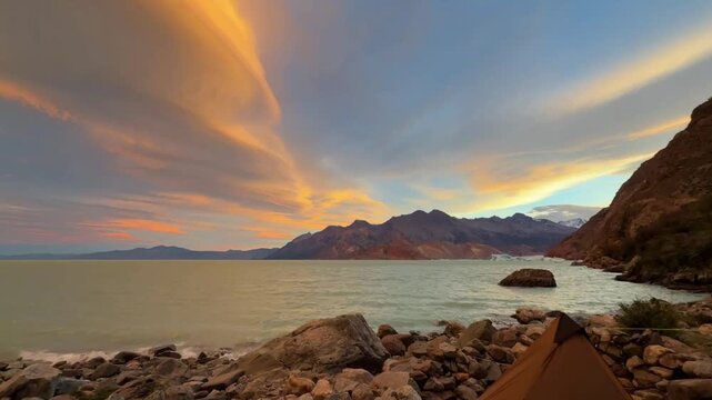 Expedition basecamp in Patagonia: Camping tent on a stony glacier lake shore facing majestic mountains under a colorful twilight sunset sky.