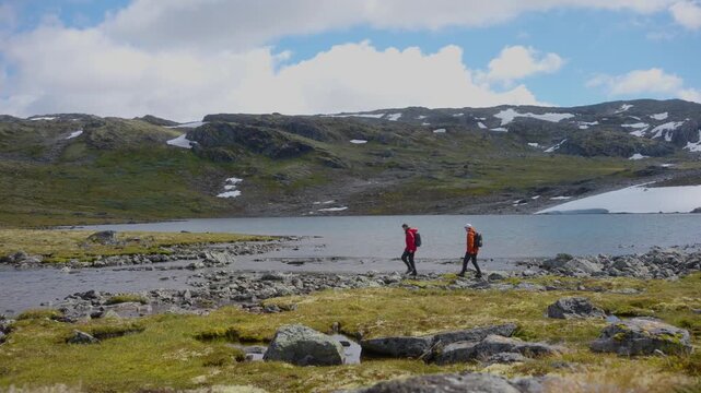 Two hikers cross a small mountain stream flowing from Flyvotni in the highlands of Norway. The rugged alpine landscape features rocky terrain, low vegetation, patches of lingering snow and mountain 
