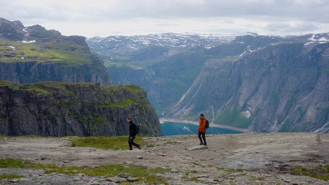 Two hikers walk toward Trolltunga through the dramatic mountain landscape of western Norway. Steep cliffs, deep valleys, a mountain lake and snow-covered peaks create a spectacular alpine backdrop. St