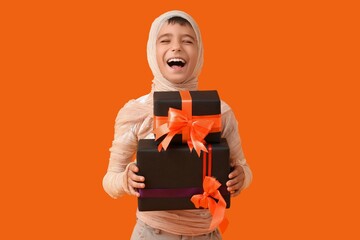 Obraz premium Happy boy in mummy costume holding halloween gifts, trick or treat celebration, and holiday party fun on orange background
