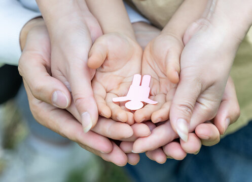 Adult and child hands cup together to hold a small pink human-shaped figure, conveying themes of care and family.