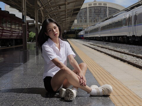 A young smiling Asian woman wearing a university student uniform sits cross-legged on a train station platform