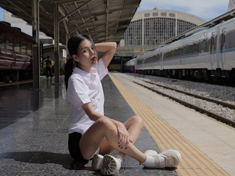 A young smiling Asian woman wearing a university student uniform sits cross-legged on a train station platform