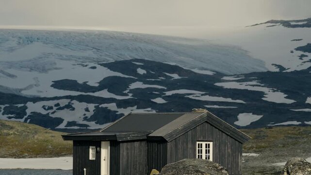 Cinematic lateral tracking shot of a traditional Norwegian mountain cabin surrounded by rugged alpine scenery on Sognefjellet. The dark wooden hytte stands in the foreground while the vast B&oslash;verbreen 
