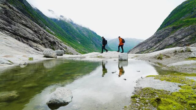 Two hikers walk across smooth glacial rock beside a small mountain pool in the Nigardsbreen area of Norway. The calm water reflects the hikers and surrounding steep green-covered mountains, creating a