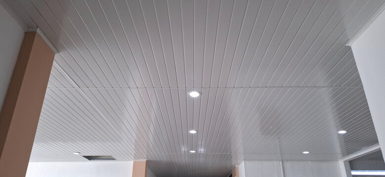 Low angle perspective shot of a modern white PVC striped ceiling featuring recessed LED downlights and structural side pillars. Ideal for interior construction, ceiling material samples, and modern de