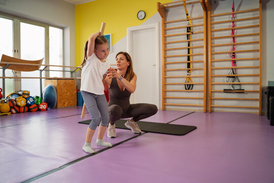 A fitness instructor guides a young girl's posture and technique as she performs a mobility exercise with a wooden stick behind her back in a gym