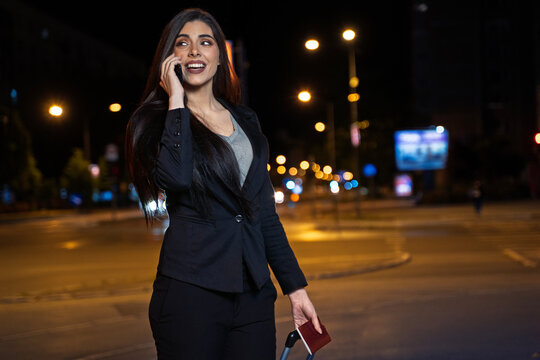 A smiling young woman in a black blazer talks on her phone and holds a passport while walking with a suitcase at night.