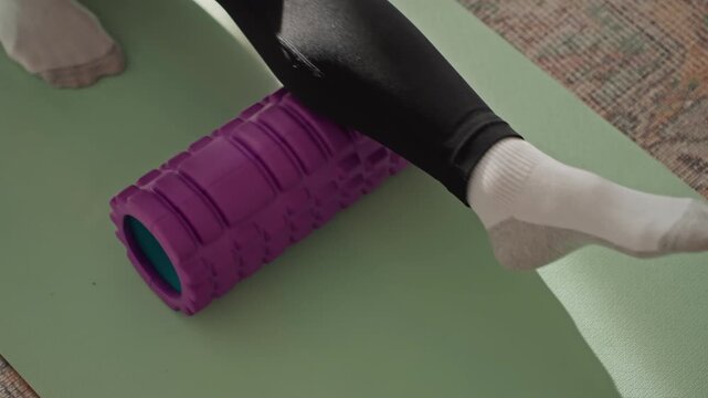 Close up white woman foam calf, on green mat at home, textured purple roller under lower leg, wearing black leggings and white socks, slow selfmassage movement for muscle recovery, quiet morning light