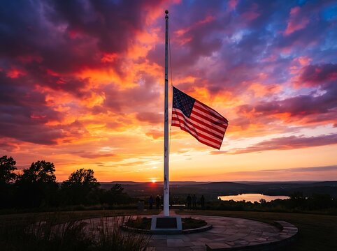 American flag flying at half mast against a dramatic and colorful sunset sky over a peaceful landscape