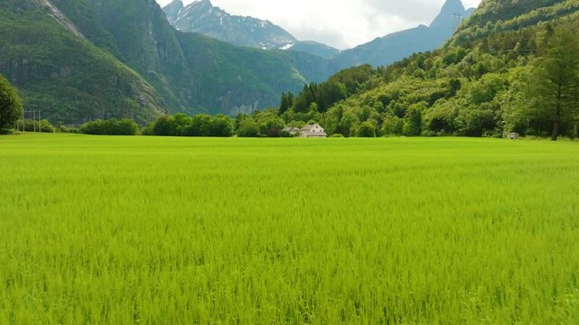 Cinematic drone footage flying low over a green field toward a traditional Norwegian farmhouse, gradually rising to reveal  dramatic Romsdalen valley and surrounding mountains near &Aring;ndalsnes, Norway