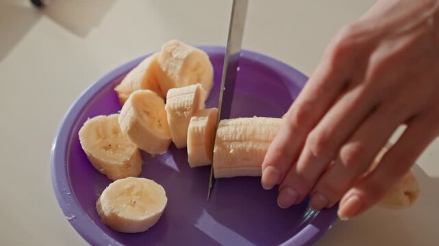 Closeup hands chopping banana into rounds on purple plate, dynamic cutting motion, scattered slices, watch on wrist, quick home snack prep for fitness, energetic morning mood, tactile focus, serving