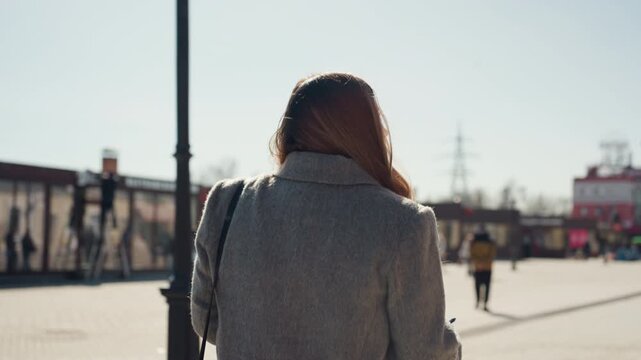 Woman strolling outdoors. Lady with hair moving in sunlight. Female figure wearing overcoat walking through cityscape. Woman with shoulder bag strolling in lively public square during daytime