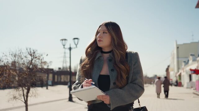 Pretty woman walking city plaza carrying notebook and pen, thoughtful gaze toward red brick building, sunny afternoon, lamp posts and sparse pedestrians, cinematic shallow depth, casual jacket