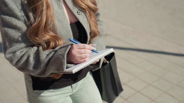 Entrepreneur outlining plans on city street. Woman entrepreneur writing in notebook on busy sidewalk. Confident female startup founder drafting her strategy in urban environment with sunlight