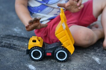 Child playing outdoors with a yellow toy dump truck on pavement. Close-up of hands and toy vehicle, capturing creative childhood play, imagination and summer leisure activity. © Karolina