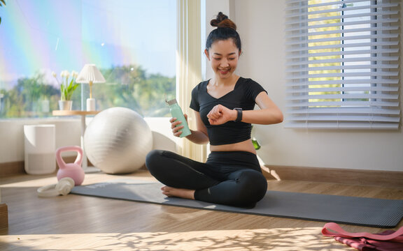 Young Asian woman checking her smartwatch and drinking water after a home workout.