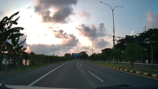 Front dashcam driving through a tropical Indonesian road beneath a dramatic morning sky with sunlight glowing behind clouds.