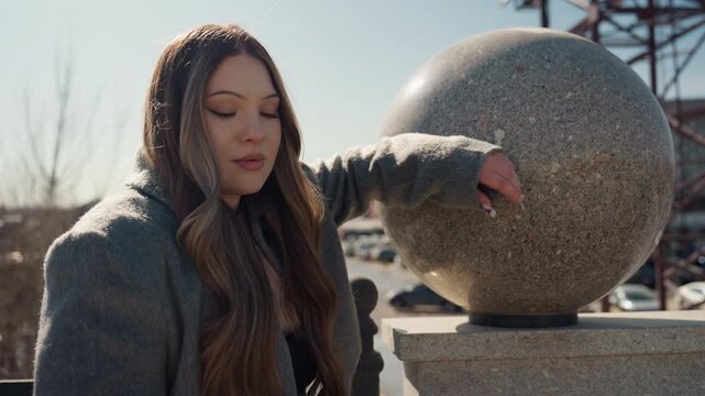 Pretty asian woman leaning on stone sphere, wool coat, long hair cascading, soft sunlit winter light, distant train station and cars blurred, contemplative gaze toward horizon, cinematic warm tones,