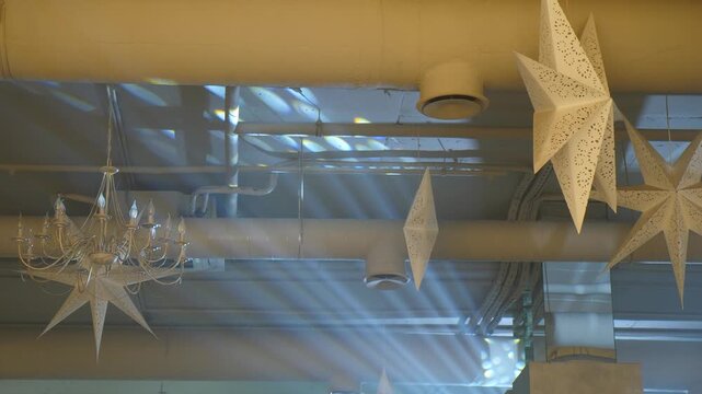 Close-up of industrial pipe ceiling with hanging white paper stars and crystal chandelier, dramatic sunbeams streaming through windows creating unique atmospheric dance club party space 4k video