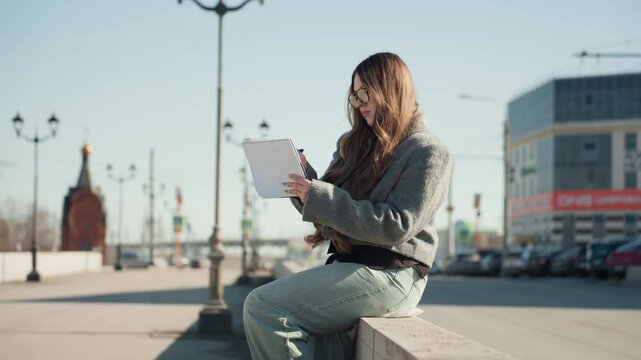 Pretty caucasian woman journaling on bench soft morning light, relaxed posture, gray coat, glasses, pages filled with handwriting, distant lampposts and quiet plaza, reflective mood