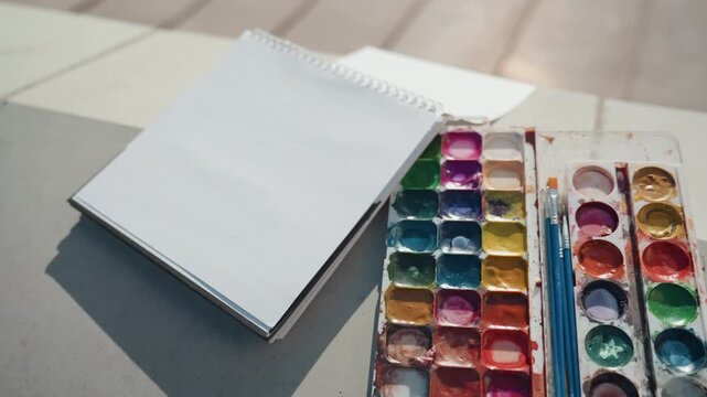 Waiting for colorful artwork creation. Enjoying peaceful hours with watercolors and sunshine. Engaging in calming artistic pursuits using watercolors on textured surfaces during sunny weather