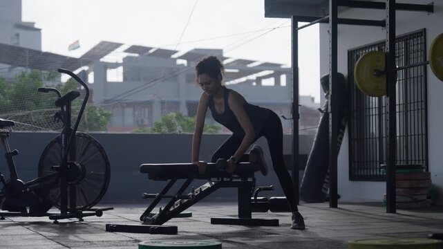South Asian female athlete performing single-arm dumbbell rows supported on a bench at an open-air rooftop gym. Full shot with static camera angle. Collection: WORKOUTSERIES2026.