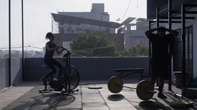 South Asian male athlete performs barbell power cleans while a female athlete trains on an assault air bike at an open-air rooftop gym. Wide tracking shot. Collection: WORKOUTSERIES2026.