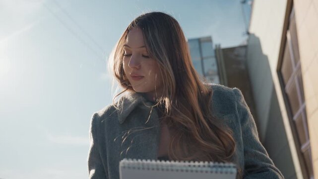 Pretty asian student sketching outdoors, lowangle sunlit city scene with wool coat and notebook, thoughtful downward gaze while turning pages, cinematic backlight and soft shadow, gentle creative