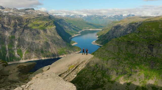 Aerial drone footage of two hikers approaching and then standing on Trolltunga, the iconic rock formation overlooking Ringedalsvatnet near Odda, Norway. The sequence shows the hikers walking toward 