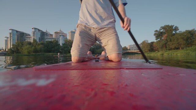 Man Kneeling on Paddle Board Rowing on River | Ultra Wide-Angle Low POV Shot with Paddle and City Skyline | Sunset Water Sport Recreation Lifestyle